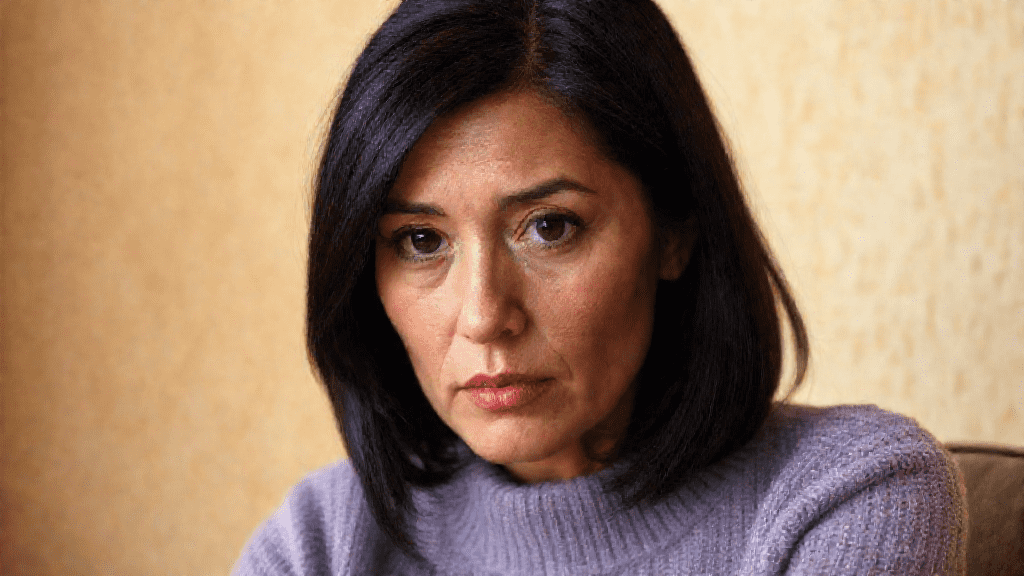 A woman with shoulder-length black hair and a serious expression is looking directly at the camera, wearing a light purple sweater, against a beige background.